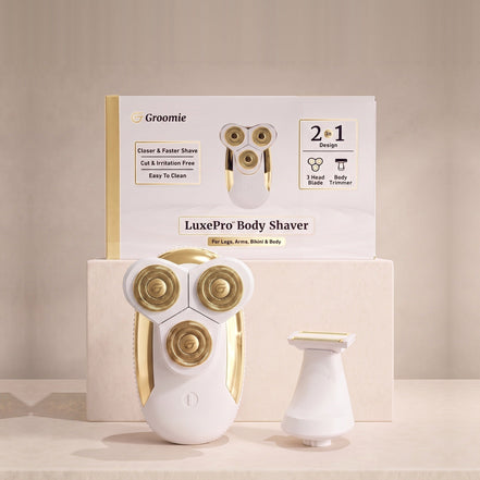 LuxePro Women's Body Shaver, a white and champagne gold rotary shaver, shown with its Groomie box on a warm cream background.